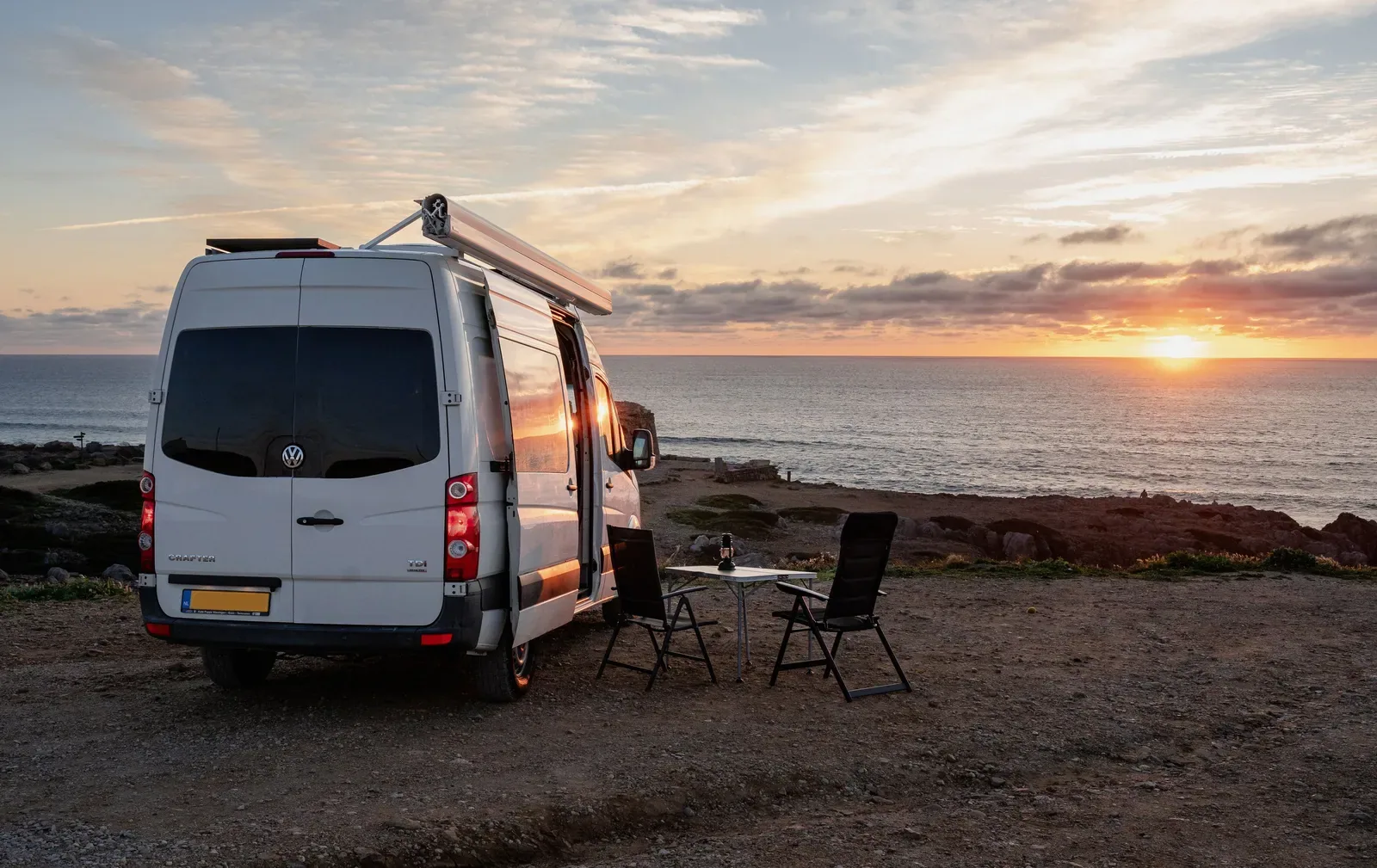 Off-grid buscamper Horizon met uitgerolde luifel, tafel en stoelen aan de Atlantische kust bij zonsondergang